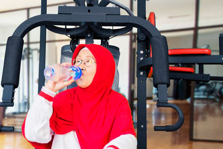 Portrait of senior muslim woman drinking a bottle of water while leaning on a boxing bag, isolated...