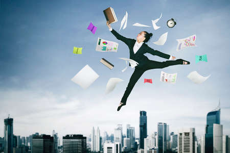Young businesswoman reading a book while jumping with documents in the blue sky over the modern cityの写真素材