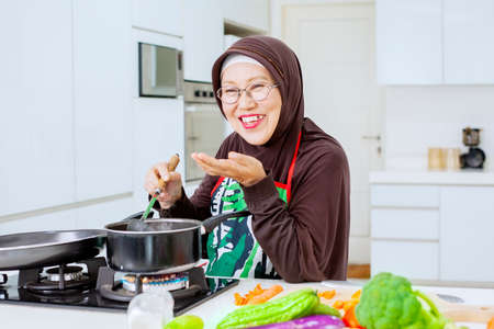 Happy old woman tasting soup from a pot while standing in the kitchenの写真素材