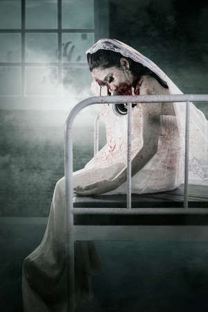 Halloween horror concept. Horrible female bride ghost with wounded face sitting on the bed with spooky night view background on the windowの写真素材