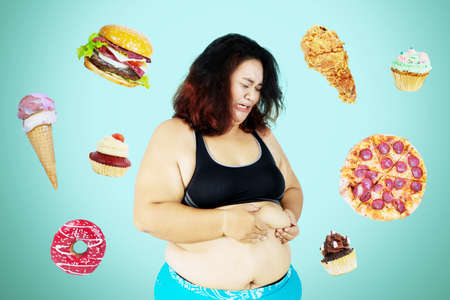 Portrait of obese woman looks cry while pinching her belly fat and standing with junk foods background. Diet fail conceptの写真素材