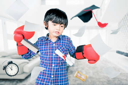 Angry little boy throwing papers and graduation cap while wearing boxing gloves in the bedroom at...
