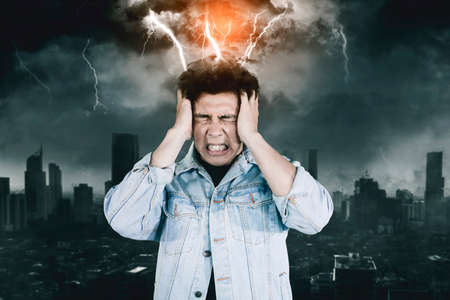 Close up of stressed young man suffering headache with attacking lightning his head with storm city backgroundの写真素材