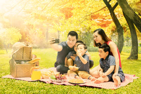 Happy family picnicking together in the park while using a mobile phone to taking selfie picture with autumn trees backgroundの写真素材