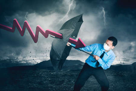 Young businessman attacked by declining arrow while holding an umbrella with stormy sky background