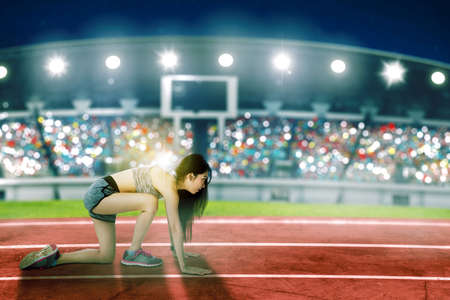 Side view of a female runner getting ready to start the race while kneeling on the track line with crowded spectators in the stadiumの写真素材