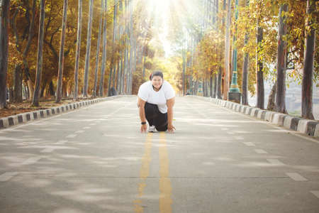 Young overweight woman smiling at the camera while ready to run and kneeling on the road at autumn time. Shot at the parkの写真素材