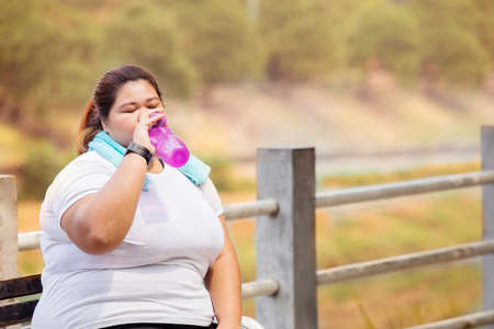 Overweight woman drinking a bottle of water after doing run exercises while sitting in the park with autumn leaves backgroundの写真素材