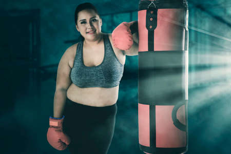 Fat woman smiling at the camera while standing near a boxing sack after exercising in fitness centerの写真素材