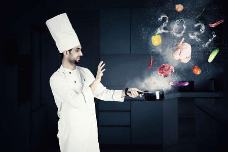 Male magician chef cooking on a saucepan with flying food ingredients and smoke shaped of numbers 2021 in the kitchenの写真素材
