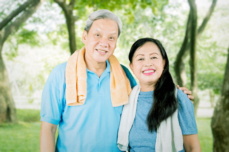 Elderly couple smiling at the camera while wearing sportswear and standing in the park with trees backgroundの写真素材