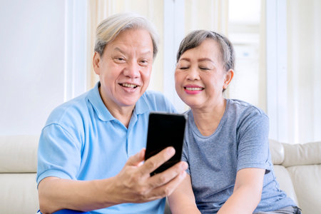 Happy old couple making a video call by using a cellphone while sitting on the sofa. Shot at homeの写真素材