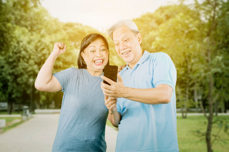 Happy old couple reading good news on cellphone while standing at the park with sunlight backgroundの写真素材