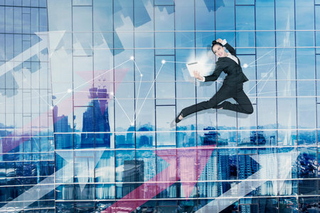 Happy businesswoman using a sparkling tablet while dancing with upward arrows and skyscraper windows reflected modern cityの写真素材