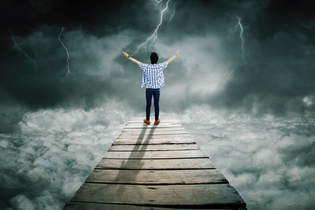 Rear view of young man looks bravery while standing on the wooden bridge and looking at lightning at cloudy skyの写真素材