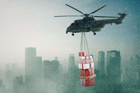 Helicopter carrying Christmas gift boxes while flying over the city at snowfallの写真素材