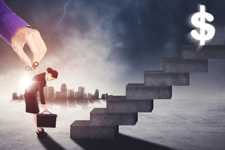 Hand giving motivation while turning winder key on tired businesswoman near stairs toward dollar symbol with cloudy sky backgroundの写真素材