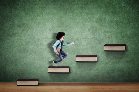 Afro male college student carrying book and bag while climbing stairs of book with chalkboard backgroundの写真素材