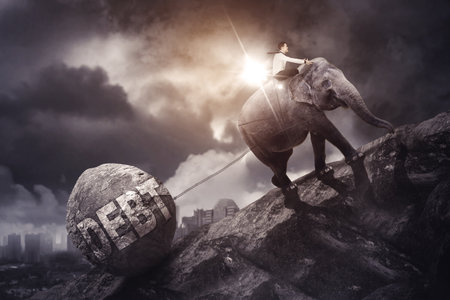 Side view of businessman riding an elephant while pulling a stone with debt word and climbing mountain with cloudy sky backgroundの写真素材