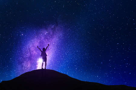 Back view of businesswoman expressing success by raising hands while standing on the hill with milky way backgroundの写真素材