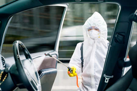 Male disinfector wearing protective suit while cleaning car interior with disinfectant spray and...