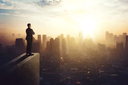 Confident businessman standing on the building rooftop while looking at the silhouette of cityscape at dusk timeの写真素材