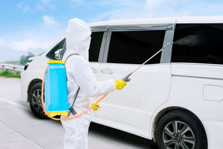 Disinfectant service worker wearing protective suit while spraying a car with disinfectant during...