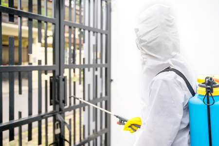 Back view of male worker wearing Hazmat suit while spraying disinfectant to house gate during...