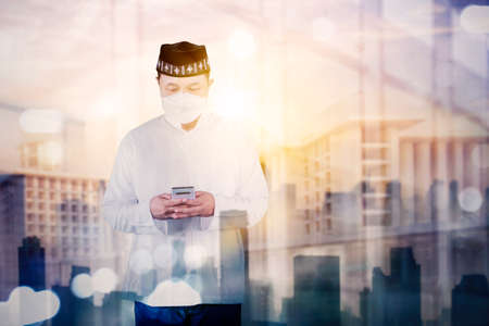 Double exposure of Muslim man wearing face mask while using a cellphone and standing with mosque backgroundの写真素材