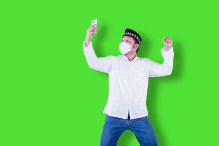 Excited Muslim man reading good news on a cellphone while wearing face mask in the studio with green...
