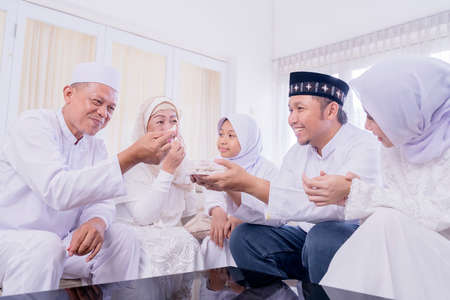 Happy Muslim young man sharing cookies to his family while eating together during Eid Mubarak in the living room at homeの写真素材