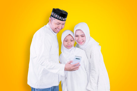 Muslim little girl with her parents making a video call by using a cellphone while celebrating Eid Mubarak in the studio with yellow backの写真素材