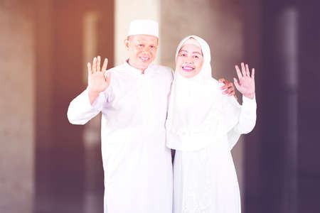 Smiling Muslim senior couple waving hands at the camera and standing together in the mosqueの写真素材