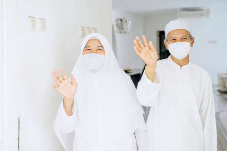 Muslim elderly couple wearing face mask while waving hands at the camera and doing quarantine at home during Eid Mubarakの写真素材