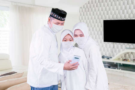 Happy parents with their daughter wearing face mask while making a video call by using a cellphone...