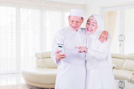 Smiling Muslim old couple using a mobile phone while standing together in the living room at homeの写真素材