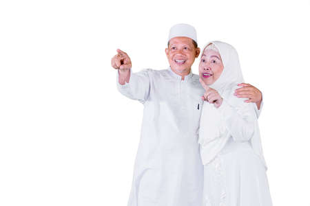 Happy Muslim old man embracing his wife while looking and pointing at copy space in the studio. Isolated on white backgroundの写真素材