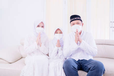 Happy Muslim family wearing face mask while showing congratulate hands gesture Eid Mubarak and sitting on the sofa. Shot at homeの写真素材