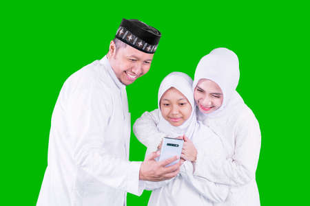Happy parents with their daughter making a video call by using a cellphone while celebrating Eid Mubarak in the studio with green screen backの写真素材