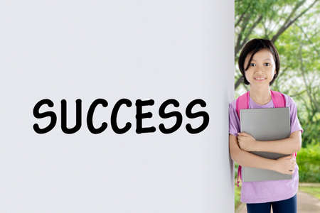 Image of adorable schoolgirl leaning on the wall with success word while holding a laptop. Shot at outdoorの写真素材