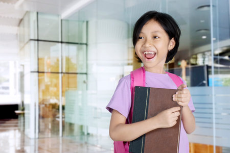 Picture of adorable schoolgirl holding a book with happy expression. Shot in the schoolの写真素材