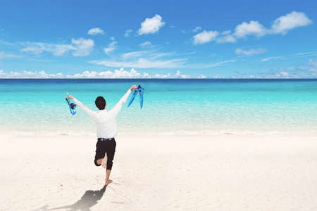 Back view of happy businessman running on the beach while carrying snorkeling equipmentsの写真素材