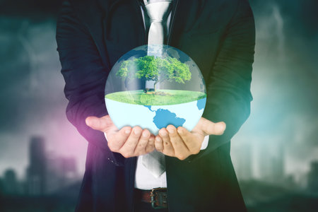 Earth Day concept. Close up of businessman hands holding an earth planet and green tree with blue sky backgroundの写真素材
