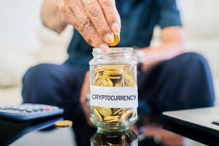Cryptocurrency investment concept. Close up of elderly man hand saving gold coins in a glass jar with Cryptocurrency word.の写真素材