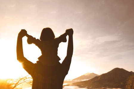 Rear view of young man doing piggyback with his daughter while enjoying sunset scenery and standing on the mountainの写真素材