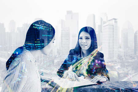 Double exposure of female manager showing a business contract document to her partner while sitting with glowing city backgroundの写真素材