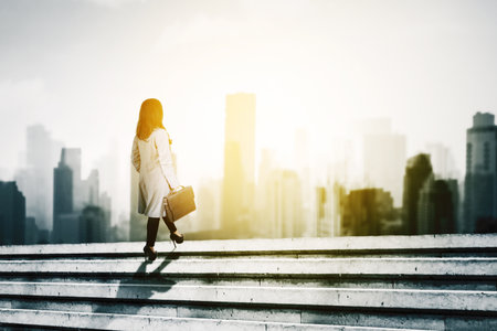 Rear view of young businesswoman carrying a suitcase while climbing stairs toward modern city with sunlightの写真素材