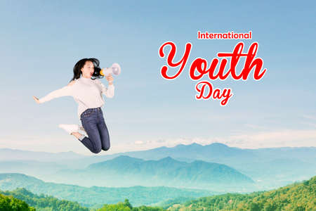 Happy young woman screaming through a megaphone while jumping with International Happy youth text in misty mountain backgroundの写真素材