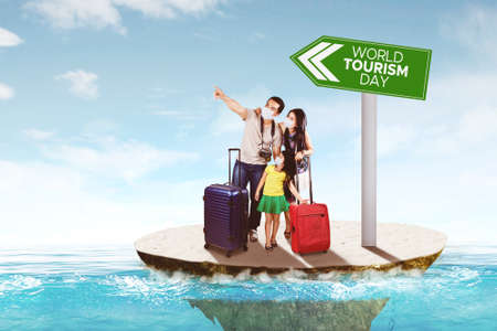 Happy young family wearing face mask while pointing at something and standing with world tourism day text on a signboardの写真素材