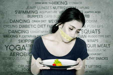 Young woman holding a plate of salad and covering her mouth with a measure tape while suffering anorexia and sitting with her problems backgroundの写真素材